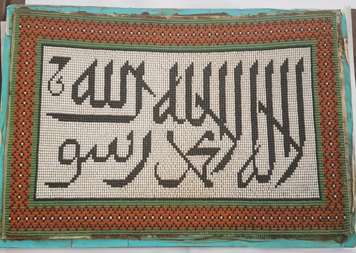 Handmade bead artwork showing two lines of black Arabic calligraphy on an ivory field, inside a terracotta-orange border patterned with black and white dots.