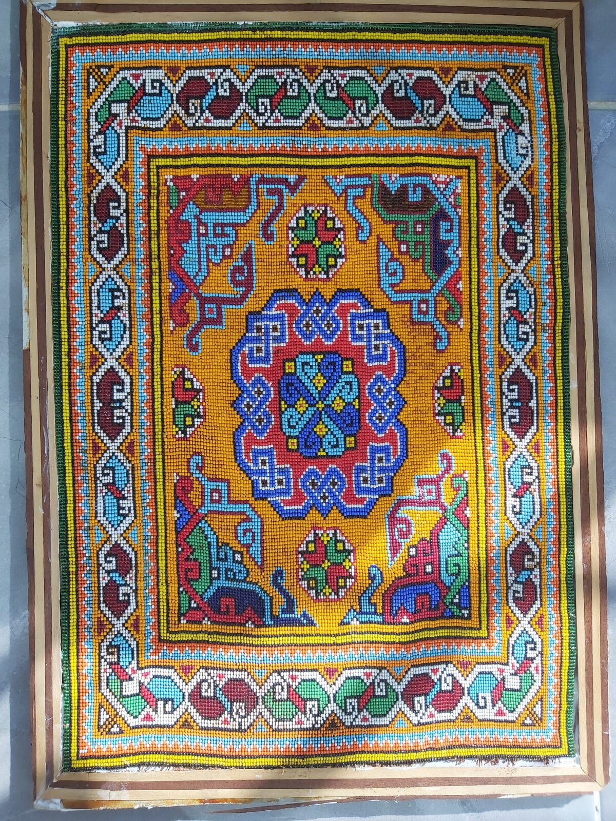 Handmade bead artwork in a striped wooden frame: a blue and red geometric medallion of hooked motifs on a saffron-ochre field, inside a white interlaced border with turquoise, burgundy and green motifs.