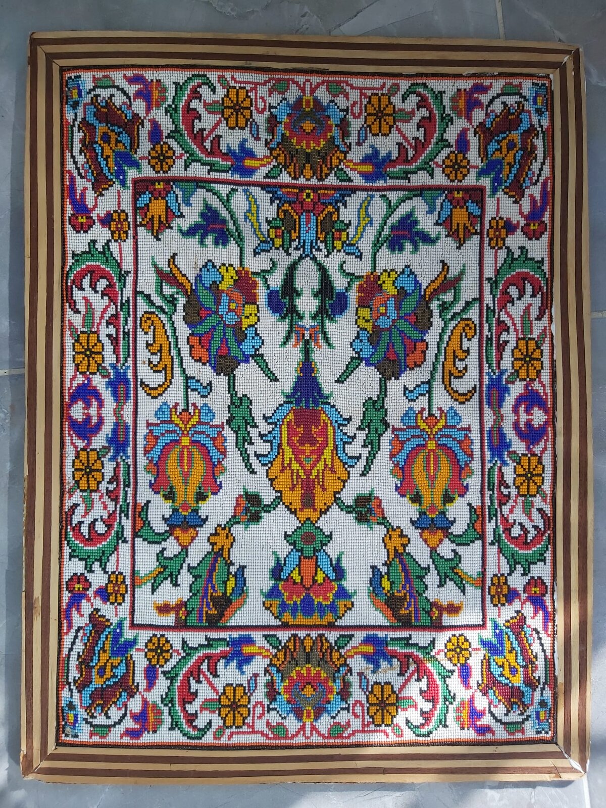 Handmade bead artwork in a striped wooden frame: multicoloured flowers on curving green vines around a golden central pendant on a white field, inside a white border of red and green scrolls.
